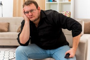 גבר בשנות השלושים לחייו, עייף, שמנמן, יושב מדוכא על ספה ושלט טלוויזיה בידו